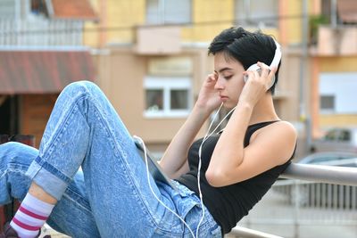Side view of young woman listening music while sitting against building in city