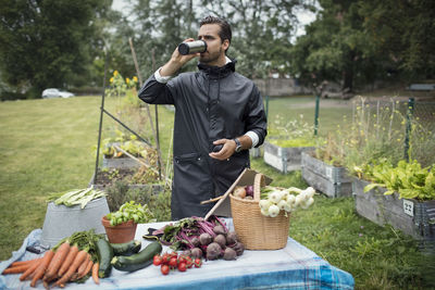 Mid adult man drinking coffee while standing by freshly harvested vegetables on table at urban garden