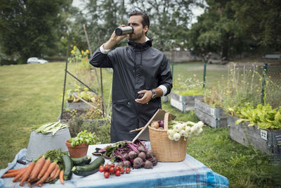 Mid adult man drinking coffee while standing by freshly harvested vegetables on table at urban garden