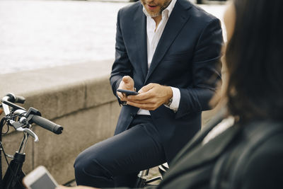 Midsection of businessman using mobile phone while sitting on bicycle by businesswoman