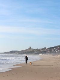 Woman running with dog at beach against sky