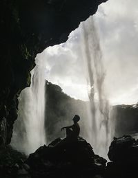 Side view of man sitting on rock by waterfall against cloudy sky