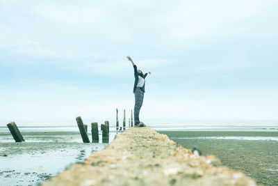 Woman with arms raised standing on jetty against sky