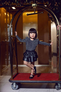 Beautiful korean girl child in a black dress is sitting on a luggage trolley in a hotel