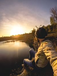 Rear view of man sitting at riverbank during sunset