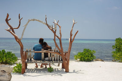 Rear view of couple sitting on bench at beach against clear sky