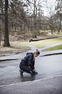 Full length side view of female athlete tying shoelace while crouching on road in forest