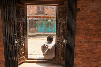 Rear view of man sitting on entrance against old house