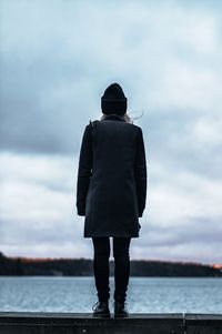 Rear view of young woman in warm clothing standing on retaining wall by lake against cloudy sky