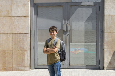 Portrait of smiling boy with backpack standing against metal door during sunny day