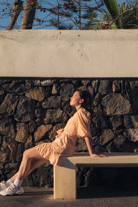 Side view of woman sitting on bench against wall