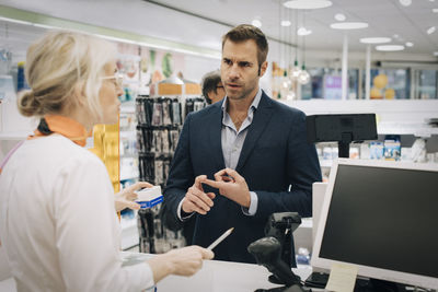 Mature man explaining to female pharmacist standing at checkout in medical store