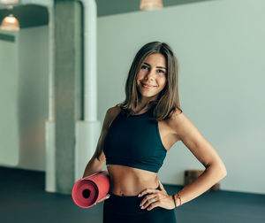 Portrait of smiling woman with exercise mat standing against wall in gym