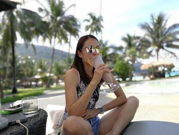 Young woman having drink while sitting against palm trees at beach