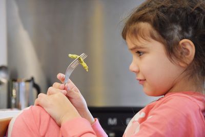 Side view of cute girl holding food with fork while sitting at home