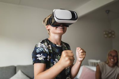 Smiling boy wearing virtual reality headset while mother sitting in background at home