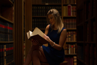 Woman reading book while sitting on stool in library