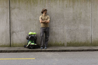 Man standing with backpack on footpath against wall