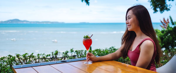 Young woman holding ice cream by sea against sky