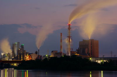 Illuminated factory by river against sky