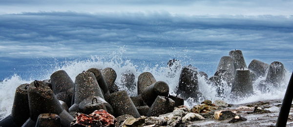 Scenic view of sea waves splashing on tetrapods against cloudy sky