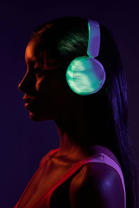 Charming brazilian female listening to music in wireless headphones while standing looking away in obscure studio on dark background with shadow of light on face