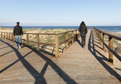 Rear view of woman and man walking on boardwalk against sea at beach