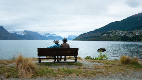 Rear view of people sitting on bench by lake against sky