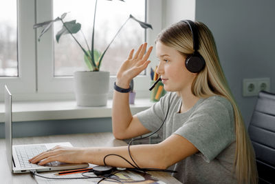 Teenage girl in headphones raises her hand while having online lesson at home