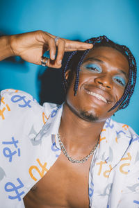 Happy african american male with bright makeup and braided hair wearing unbuttoned shirt looking at camera on blue background in studio