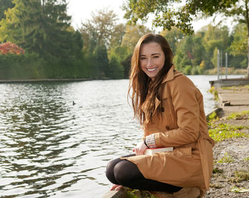 Portrait of smiling young woman crouching at lakeshore