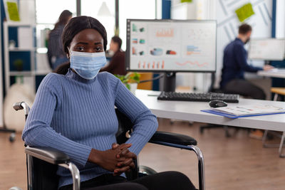 Portrait of smiling woman wearing mask sitting on wheelchair at office