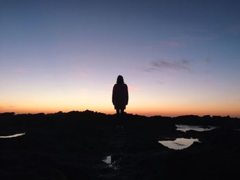 Silhouette woman standing by rocks at beach against sky during sunset