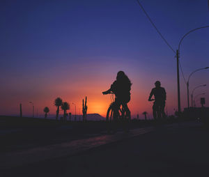 Silhouette people riding bicycle on road against sky during sunset