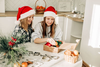Two cute girls in santa claus hats are sitting at the kitchen table and packing sweet gifts 