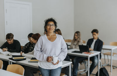 Portrait of mature female teacher holding coffee cup leaning on desk in classroom