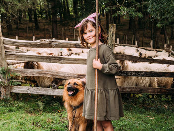 Side view of cute little girl caressing obedient basque shepherd dog near enclosure with herd of sheep in farm looking at camera