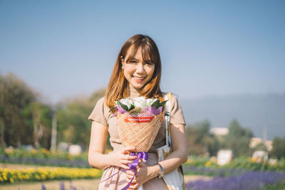 Portrait of smiling young woman holding flower bouquet while standing against sky