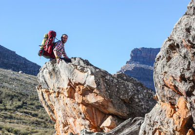 Low angle portrait of male hiker sitting on rock formation against clear blue sky