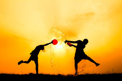 Silhouette friends splashing water on field against sky during sunset