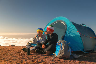 Brothers having drink while sitting on mountain against clear blue sky