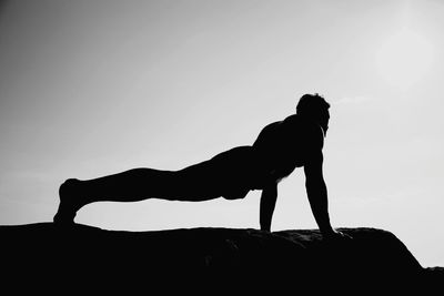 Side view of silhouette man doing push-ups on rock against clear sky