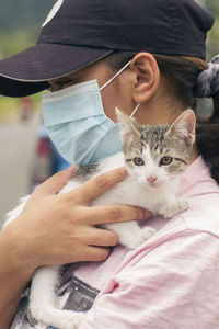 Unrecognizable woman wearing face mask and black cap carrying a young white and gray cat on her shoulders in a blurred natural environment