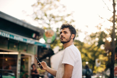 Side view of young man looking away while standing on street in city