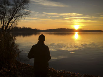 Rear view of silhouette man standing on lake against sunset sky