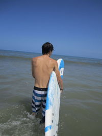 Rear view of man holding surfboard while standing in water at beach