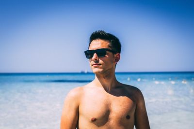 Shirtless young man looking away standing at beach against sky