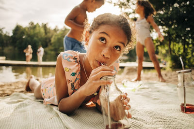 Girl looking away while drinking juice from bottle lying down on picnic blanket during vacation