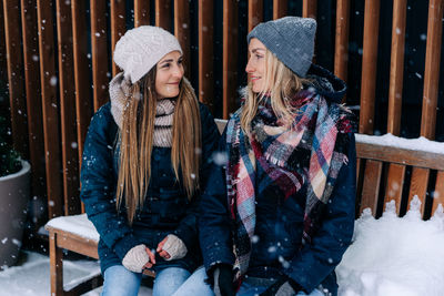 Two female friends in winter coats and hats look at each other smiling.
