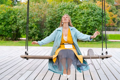 Happy woman swinging on swing in city park, wearing yellow dress and blue coat